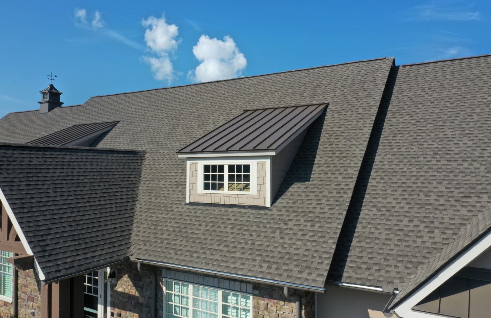 Grey asphalt shingle roof with a dormer topped in dark standing seam metal, under a blue sky.
